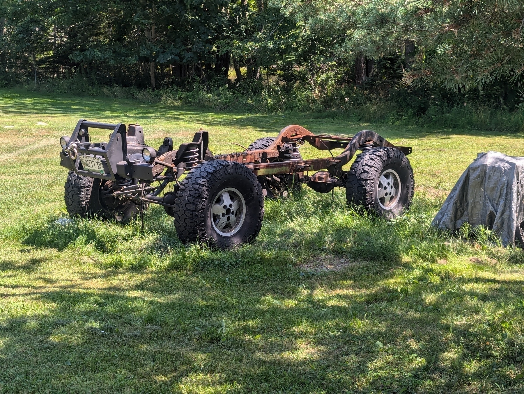 A rusty vehicle chassis with large off-road tires sits in a grassy area near a covered object, surrounded by trees.