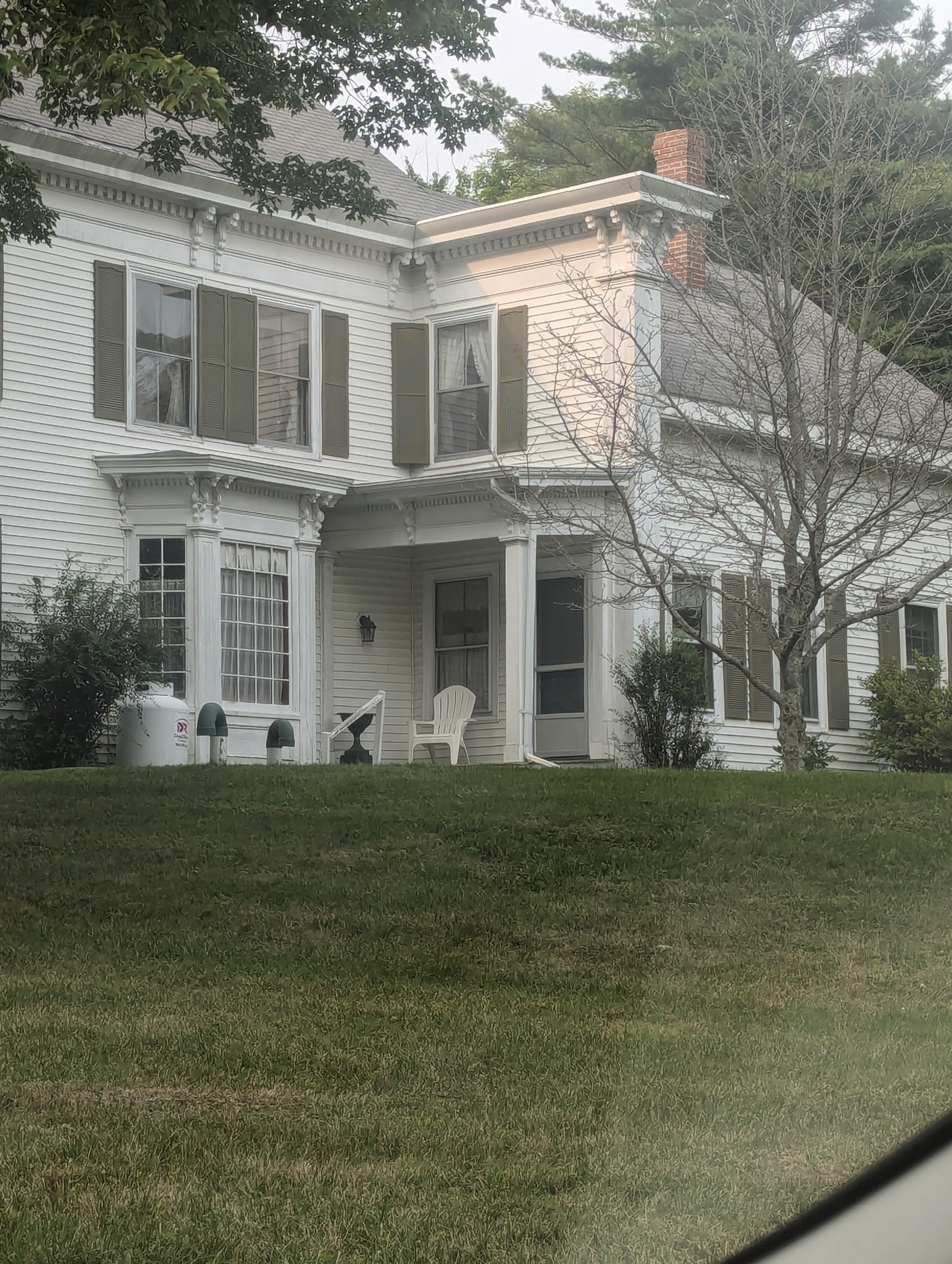 A white two-story house with green shutters features a spacious lawn, a porch with chairs, and some trees.